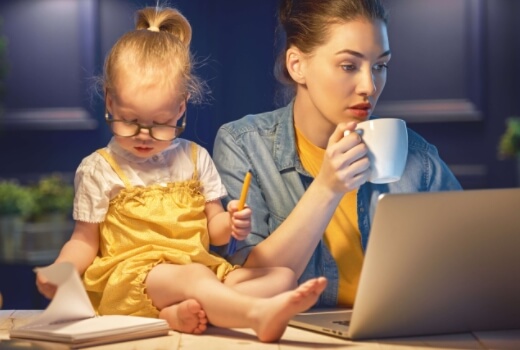 Mama dirba prie kompiuterio su kavos puodeliu rankoje, šalia ant stalo sėdi maža mergaitė, kuri įmituoja dirbančią mamą