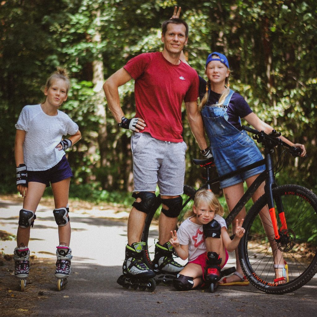 Tėtis su dviem dukromis važinėjasi riedučiais, trečioji važiuoja dviračiu.