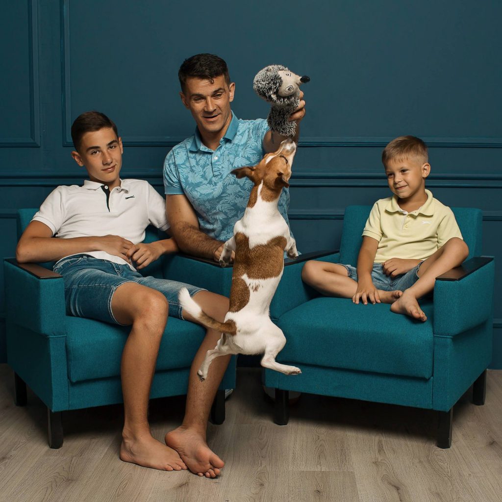 Tėtis su dviem sūnumis sėdi ant fotelių. Tetis laiko rankose žaisliuką, kurį pagriebti pašokęs šuniukas