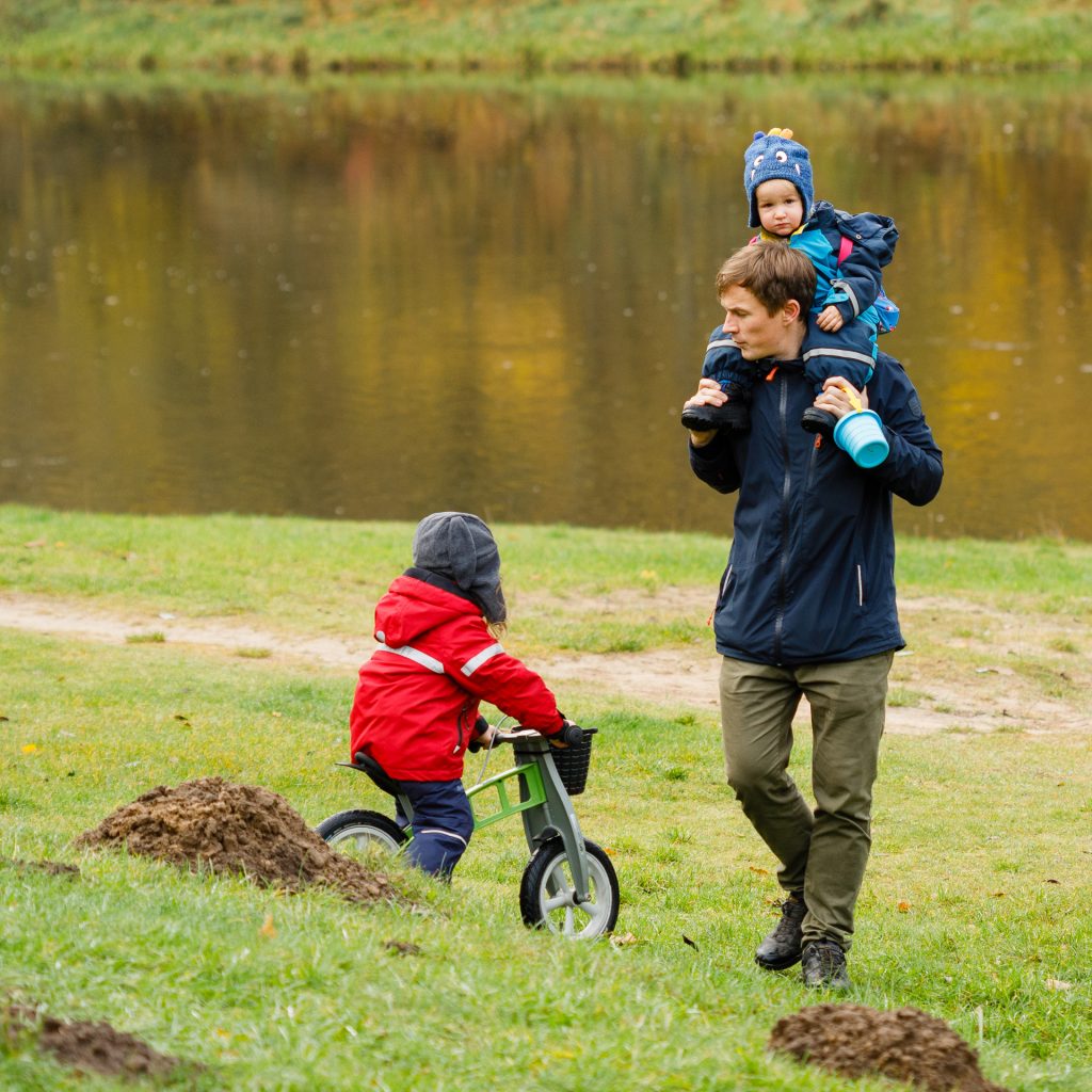Tėtis su dviem mažais vaikais gamtoje: sūnelis ant pečių, o dukra ant dviratuko.
