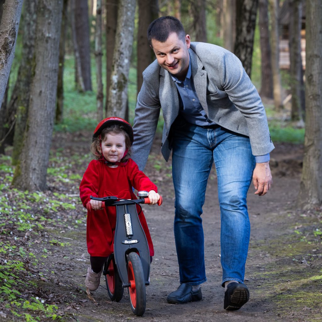 Tėtis su trimete dukrele miške, stumia ją dviračio