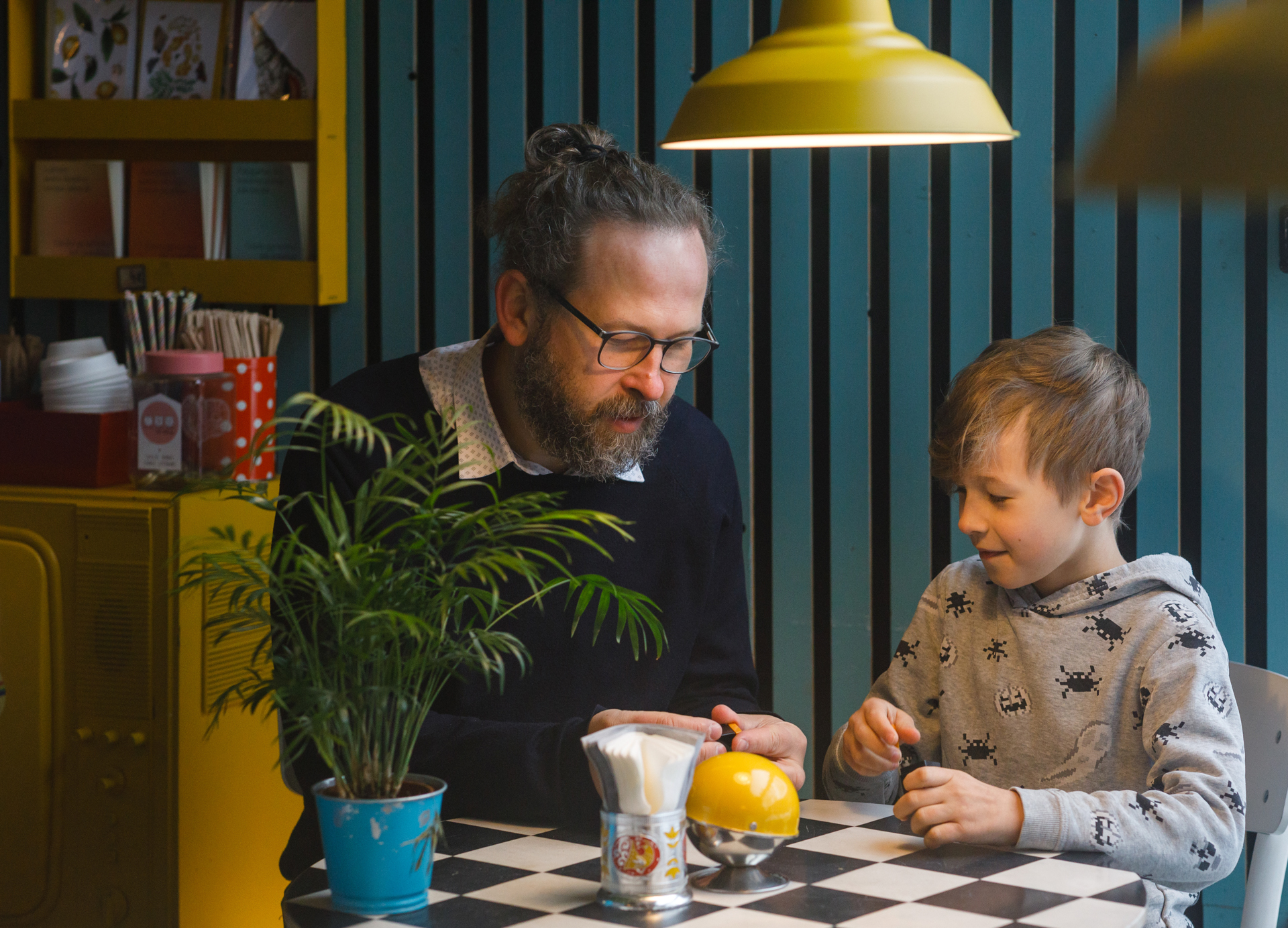 Tėtis su sūnumi kavinėje, prie staliuko sus susidomėjimu žiūri į konstruktorių tėčio rankose