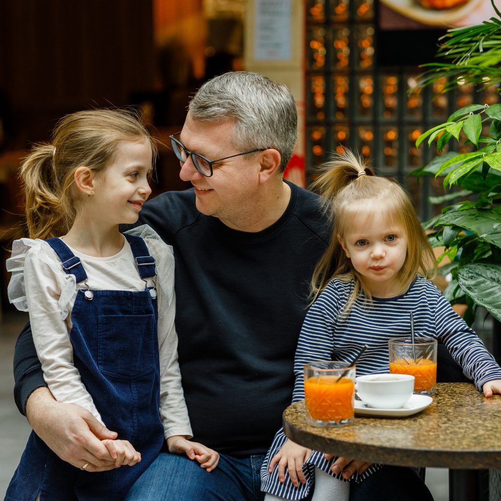 Tėtis su dviem dukrom kavinėje geria kavą ir sultis. Mažoji dukra sėdi ant kelių, didesnė prisiglaudusi iš kitos pusės.