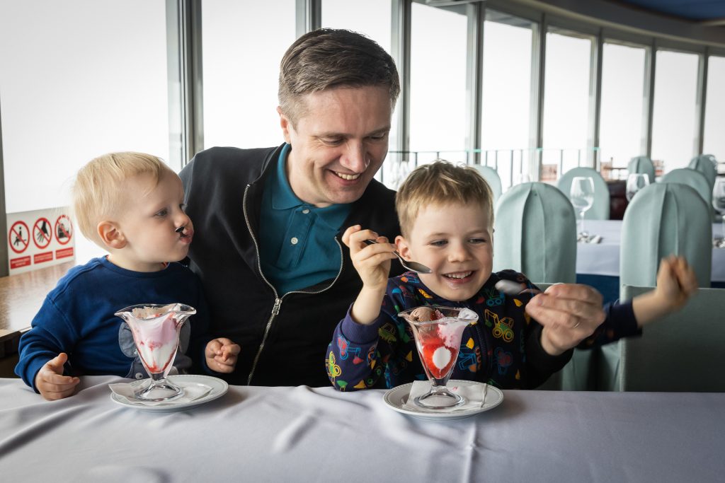 Tėtis su dviem sūnumis (penkių metų ir dviejų metų) TV bokšto kavinėje valgo ledus iš taurių.