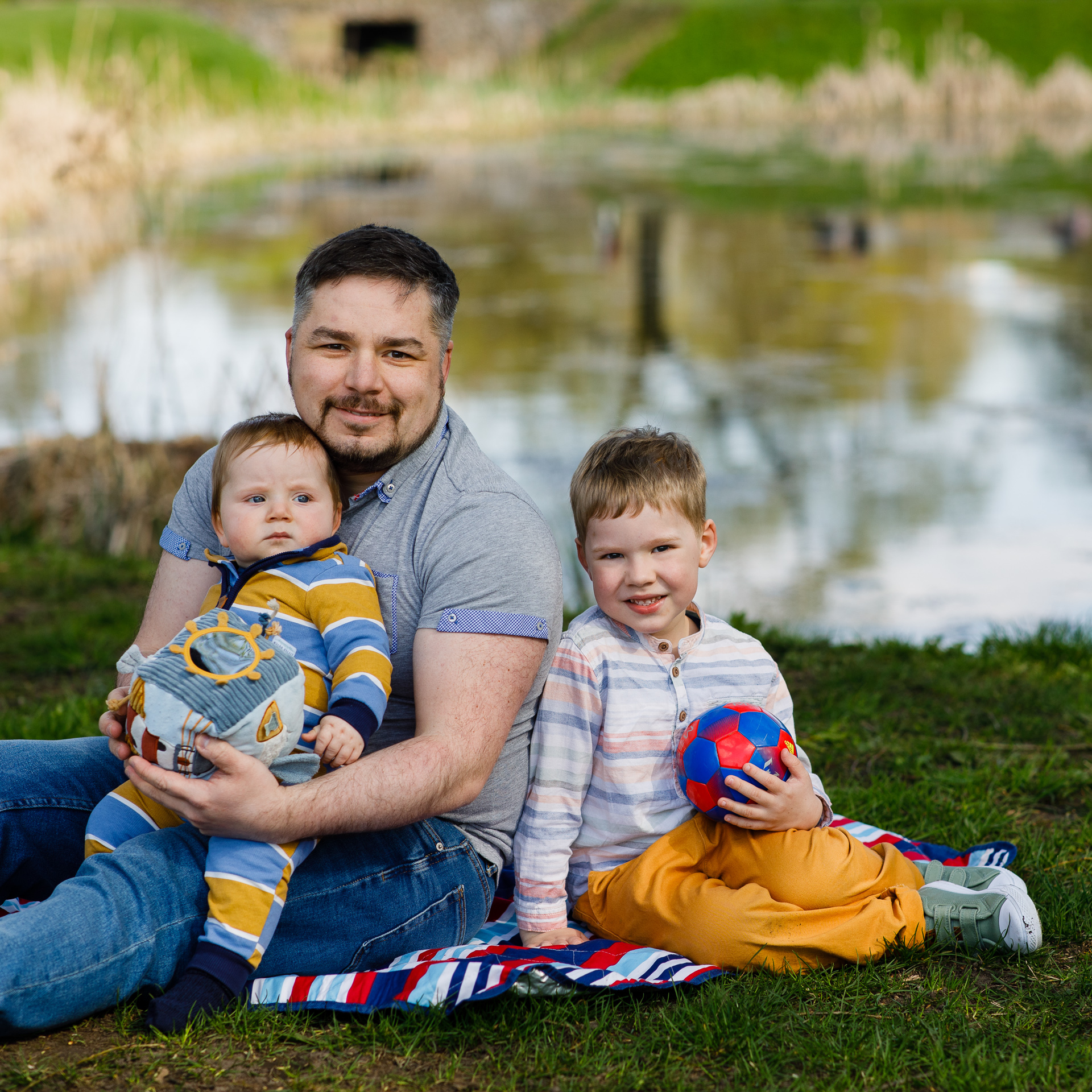Tėtis sėdo su dviem sūnumis ant pleduko prie ežero. Mažylis sunus ant rankų, šalia sėdi 4 metų berniukas. 