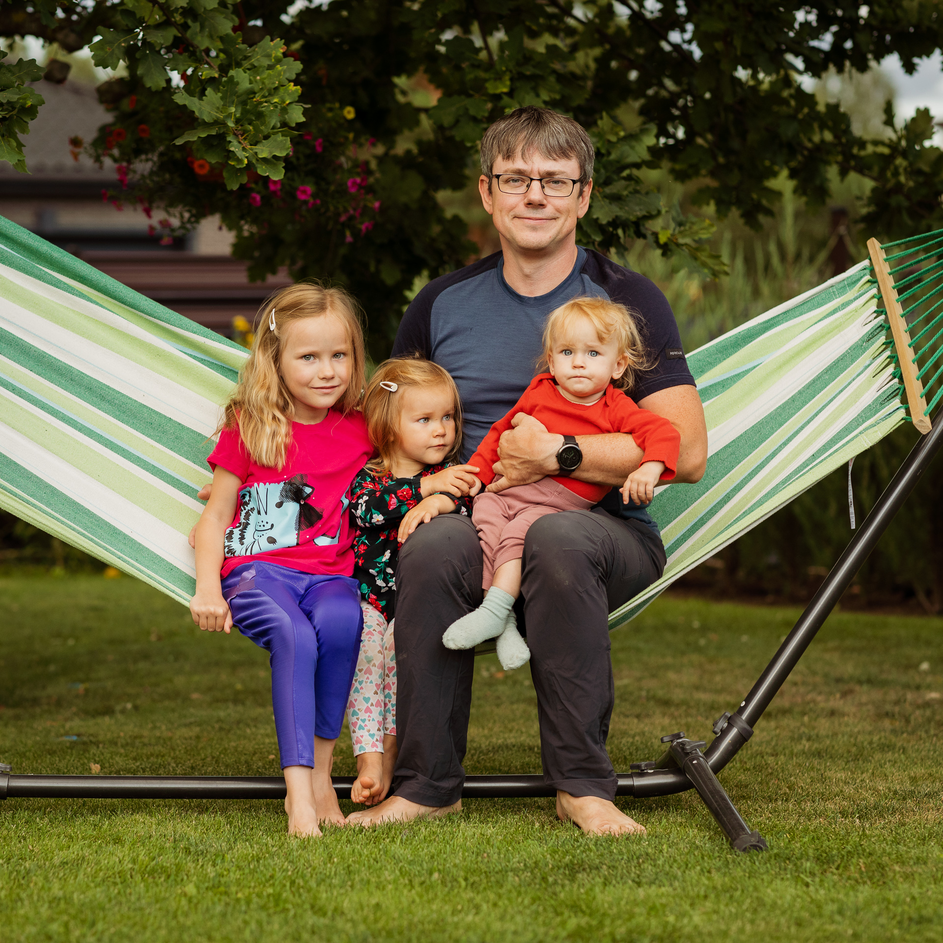 Tėtis sėdi ant hamako sode su trimis dukrelėmis. Mažiausioji ant rankų. 