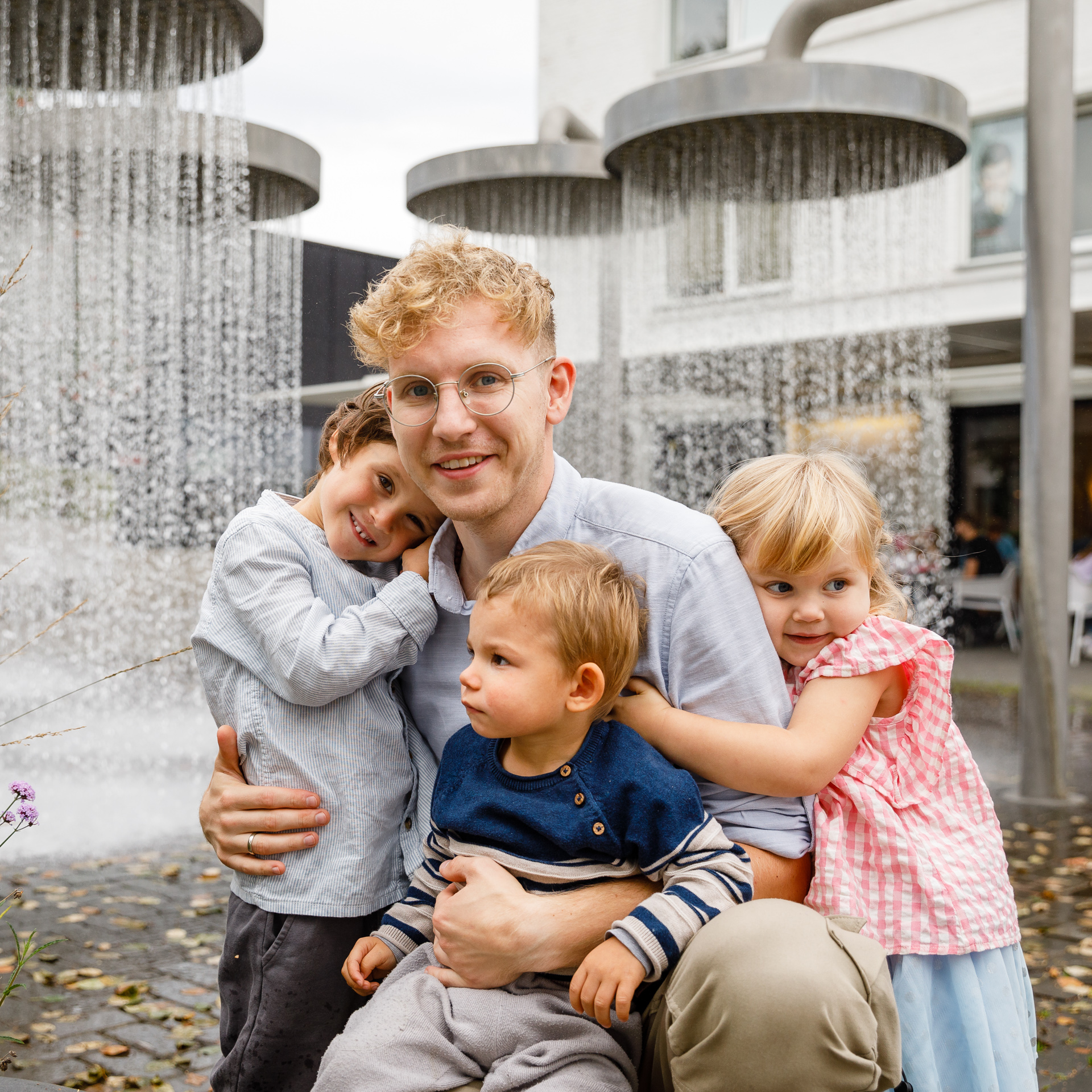 Tėtis su trim mažyliais glėbyje pozuoja prie fontano. 